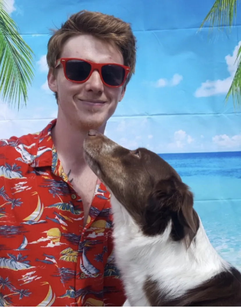 Man with glasses wearing a red hawaiian shirt with a brown and white dog looking at him.
